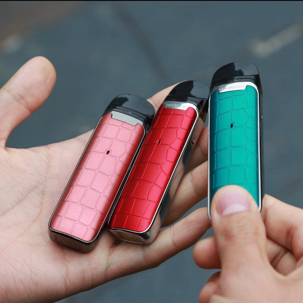 Three Vaporesso Luxe Q Pod Systems in pink, red, and teal held in hand, showcasing compact, colorful, and textured vape devices.